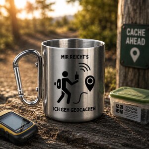 May include: A stainless steel mug with a carabiner handle, featuring a black graphic of a person with a backpack and a GPS device, along with the text "MIR REICHT'S ICH GEH GEOCACHEN" printed on the front surface of the metallic cup.