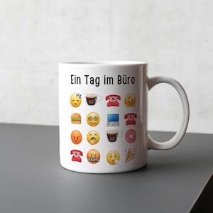 May include: White ceramic mug with a handle, decorated with various emoji icons and the German text "Ein Tag im Büro". The emojis include a sleeping face, coffee cup, telephone, and a smiling face. The mug is on a grey surface.