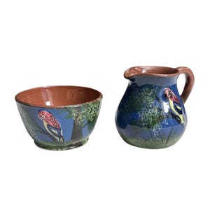 May include: A ceramic bowl and pitcher set. The bowl and pitcher are painted with a blue background and a design of a bird perched on a tree branch. The rim of the bowl and pitcher are brown.