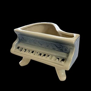 May include: A ceramic piano-shaped planter in cream, blue, and gray. The planter has a detailed keyboard design and decorative scrollwork. The piano planter is a unique decorative item.