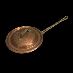 May include: A copper pot lid with a brass handle and a long brass handle. The lid is round and has a slightly domed shape. The handle is attached to the lid with two rivets.