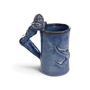 May include: A cylindrical ceramic mug with a glossy blue glaze and a dark brown rim. The handle is sculpted into the shape of a human figure leaning over the top edge. The body of the mug features a raised, embossed design of a leaf or plant stem.