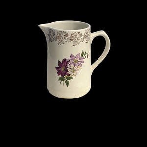 May include: Cream-colored ceramic pitcher with a floral design. The pitcher features a handle and a spout. The floral design includes purple and white flowers with green leaves. A gold floral pattern decorates the top rim.