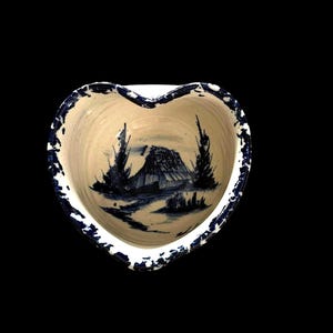 May include: A heart-shaped ceramic bowl with a cream-colored base and a dark blue hand-painted landscape scene featuring a small house and trees. The rim of the bowl is decorated with a dark blue speckled pattern against a white background.