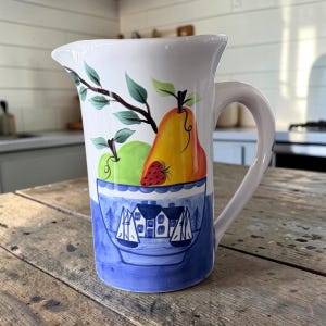 May include: A white ceramic pitcher featuring a hand-painted design of a green apple, a yellow pear, and a red strawberry above a blue and white bowl illustration. The bowl depicts a coastal scene with a house and two sailboats on the water.