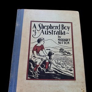 May include: A hardcover book titled A Shepherd Boy of Australia by Margaret Sutton. The cover features a black and red illustration of a youth with a dog and sheep in a landscape. The book has a light-colored front cover and a blue spine.