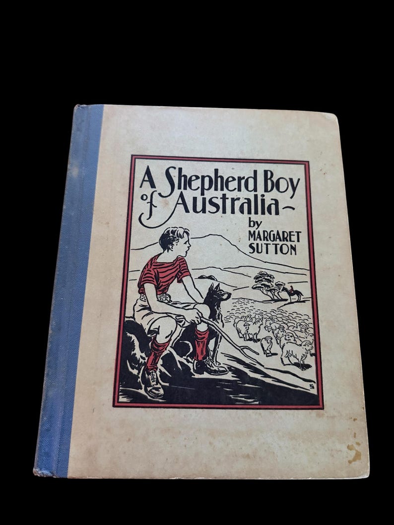 May include: A hardcover book titled A Shepherd Boy of Australia by Margaret Sutton. The cover features a black and red illustration of a youth with a dog and sheep in a landscape. The book has a light-colored front cover and a blue spine.