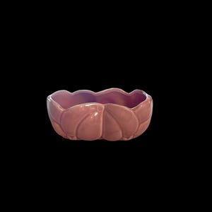May include: A pink ceramic bowl with a scalloped rim and a leaf-like design around the sides. The bowl has a glossy finish and is set against a black background. The bowl is a decorative item.