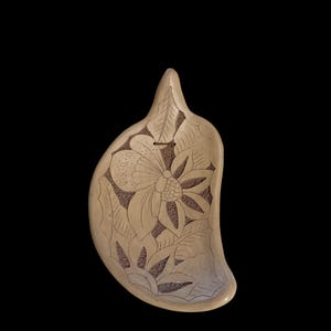 May include: A beige, leaf-shaped decorative dish with a carved floral design. The dish features a large flower in the center, surrounded by detailed leaves. The background is a darker brown, creating contrast. The dish has a smooth, curved edge.