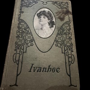 May include: A vintage hardcover book titled Ivanhoe with a sage green cloth cover. The front features a central oval portrait of a person and black line art illustrations of floral vines framing the sides. The title is printed in black serif font.