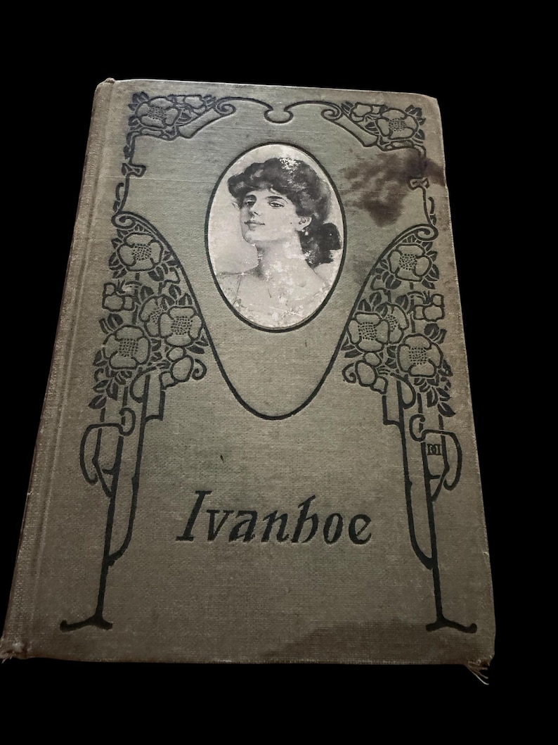 May include: A vintage hardcover book titled Ivanhoe with a sage green cloth cover. The front features a central oval portrait of a person and black line art illustrations of floral vines framing the sides. The title is printed in black serif font.