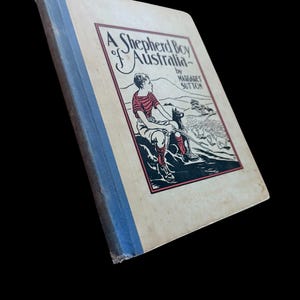 May include: A hardcover book titled A Shepherd Boy of Australia by Margaret Sutton. The cover features a cream background with a blue spine and a central illustration of a person sitting with a dog, rendered in red, black, and white ink.