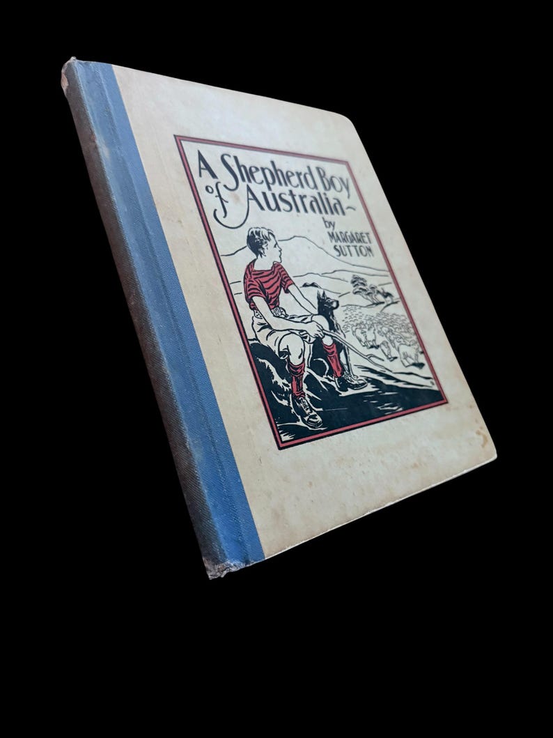 May include: A hardcover book titled A Shepherd Boy of Australia by Margaret Sutton. The cover features a cream background with a blue spine and a central illustration of a person sitting with a dog, rendered in red, black, and white ink.