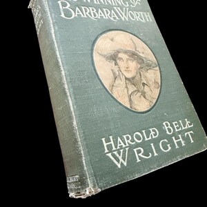 May include: A vintage hardcover book titled The Winning of Barbara Worth by Harold Bell Wright. The dark green cloth cover features white embossed lettering and a circular illustration of a person wearing a wide-brimmed hat on the front panel.