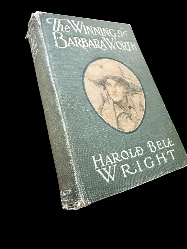 May include: A vintage hardcover book titled The Winning of Barbara Worth by Harold Bell Wright. The dark green cloth cover features white embossed lettering and a circular illustration of a person wearing a wide-brimmed hat on the front panel.