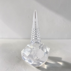 May include: A clear, faceted glass perfume bottle with a geometric base and a tall, conical stopper. The bottle's design features a series of stacked, diamond-shaped facets, creating a sparkling effect. The stopper has a ridged, textured surface.