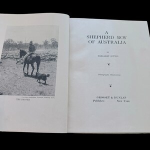 May include: An open book titled A Shepherd Boy of Australia by Margaret Sutton, published by Grosset & Dunlap, New York. The left page features a black and white photograph of a person on horseback with a dog and sheep, labeled The Drover.
