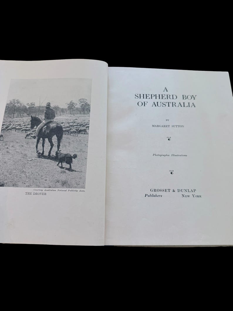 May include: An open book titled A Shepherd Boy of Australia by Margaret Sutton, published by Grosset & Dunlap, New York. The left page features a black and white photograph of a person on horseback with a dog and sheep, labeled The Drover.