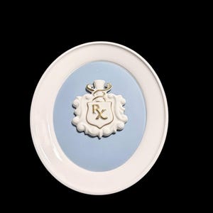 May include: A white and blue decorative plate featuring a raised emblem with the letters "Rx" in gold. The plate has a white rim and a light blue center. The emblem is white with gold accents, set against the blue background.