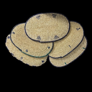 May include: A collection of five oval-shaped woven placemats. The placemats are made of natural-colored straw with dark blue and green accents. Each mat features a decorative border and geometric patterns.