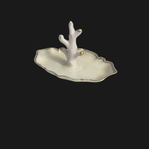 May include: A white ceramic jewelry dish with a coral-shaped accent. The dish has a scalloped edge with a gold trim. The coral accent is light pink with gold accents. The dish is on a black background.
