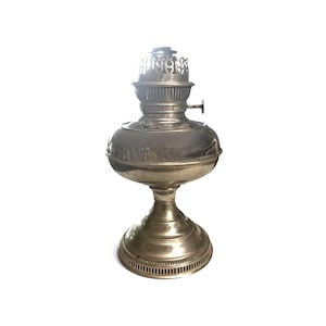 May include: A metallic oil lamp with a polished silver-toned finish. The lamp features a wide, rounded reservoir base, a slender pedestal stand with a circular foot, and a decorative burner assembly at the top with a small adjustment knob.
