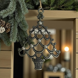 May include: A silver-colored Christmas tree ornament with pearl-like accents and the year "2004" at the base. The ornament is suspended by a natural twine string and is set against a backdrop of evergreen branches and a wooden structure.