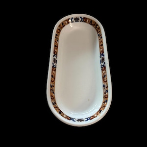 May include: An oval-shaped white ceramic dish featuring a decorative border pattern in shades of brown, blue, and gold. The interior of the dish is plain white, while the rim is adorned with a repeating intricate design of scrolls and floral motifs.