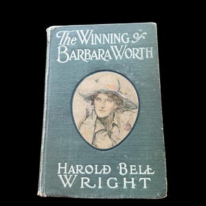 May include: A vintage hardcover book titled The Winning of Barbara Worth by Harold Bell Wright. The dark green cover features a central oval illustration of a person wearing a wide-brimmed hat, with white lettering for the title and author.
