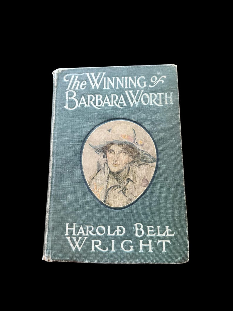 May include: A vintage hardcover book titled The Winning of Barbara Worth by Harold Bell Wright. The dark green cover features a central oval illustration of a person wearing a wide-brimmed hat, with white lettering for the title and author.