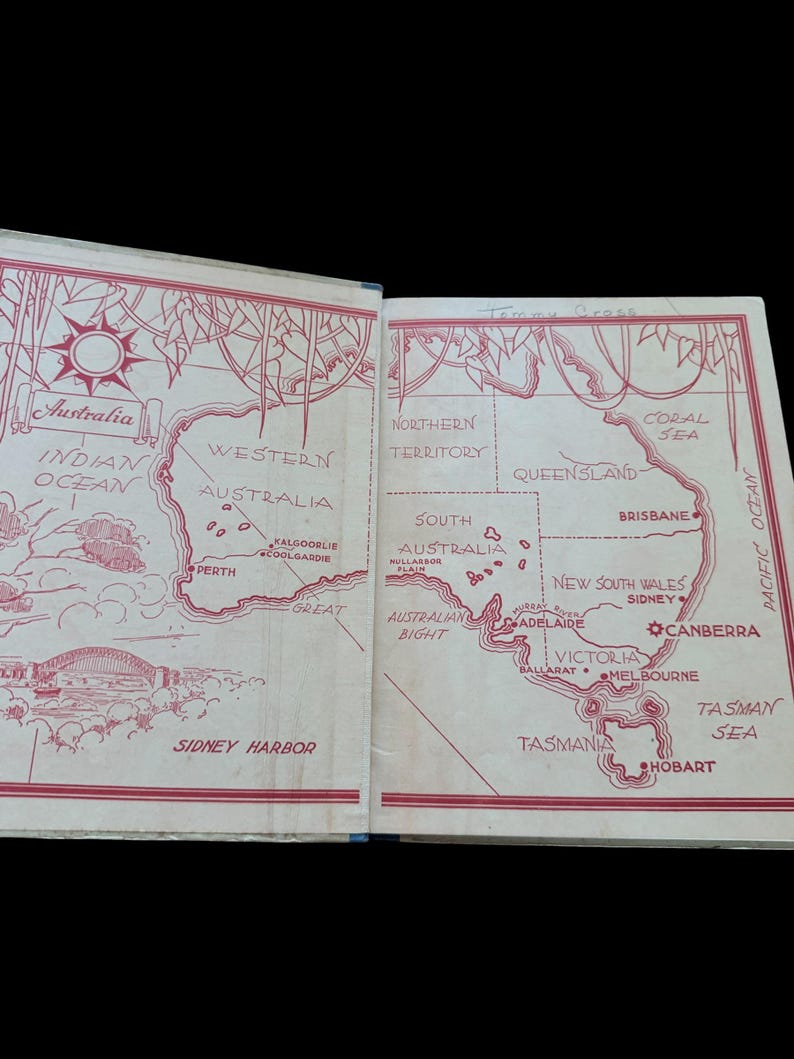 May include: An open book showing a red and white illustrated map of Australia on its endpapers. The map features labeled states, cities like Perth and Melbourne, bodies of water, and a drawing of the Sydney Harbor bridge. The text includes Australia, Indian Ocean, Pacific Ocean, and various geographic locations.