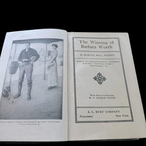 May include: An open book titled The Winning of Barbara Worth by Harold Bell Wright, published by A. L. Burt Company. The left page features a black and white illustration of a person standing near a water jar, with text below the image.