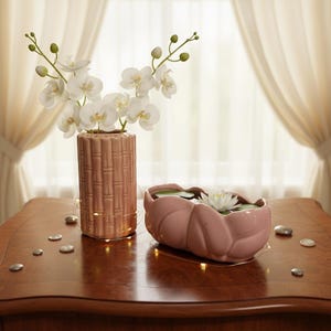 May include: A cylindrical pink ceramic vase with a bamboo-like texture containing white orchid stems, paired with an oval pink ceramic bowl holding a white water lily and green lily pads, surrounded by small silver stones and thin string lights.