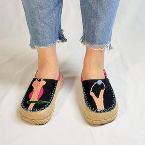 Pode incluir: Um par de sapatos pretos de lona com desenhos bordados. Um sapato apresenta um desenho de pássaro rosa e verde, enquanto o outro sapato apresenta um desenho abstrato azul e preto. Os sapatos têm uma sola de juta.