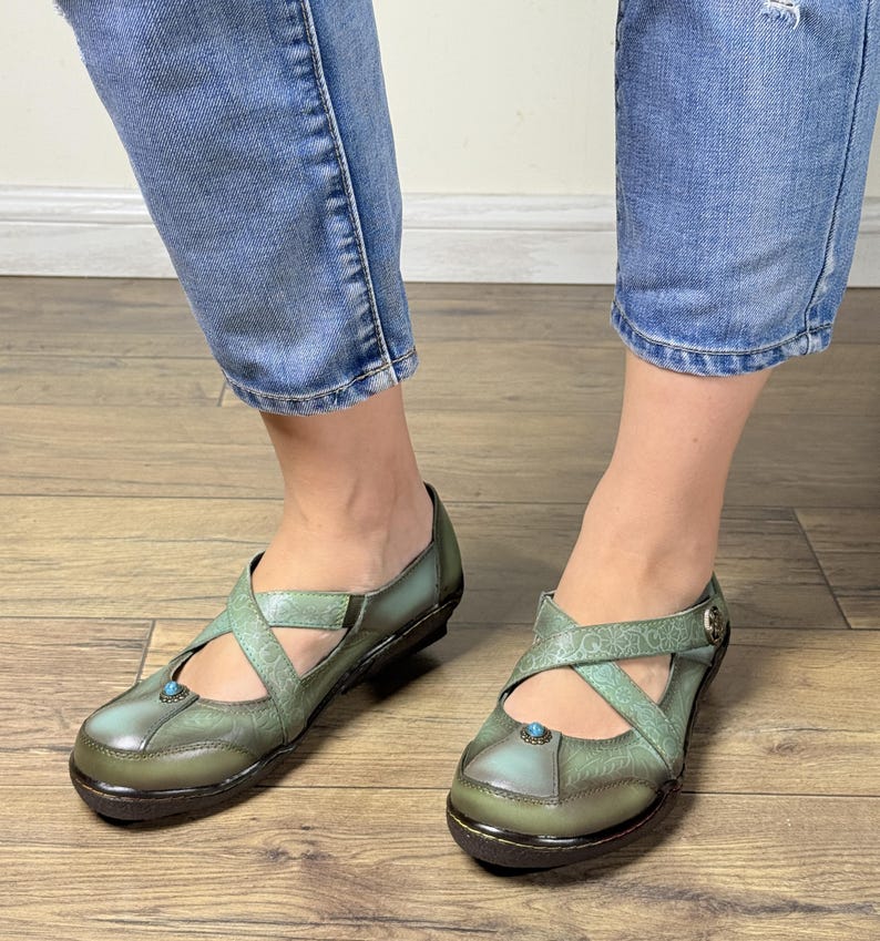May include: A pair of sage green leather shoes featuring a cross-strap design, a rounded toe, and a small turquoise stone accent on the front. The shoes have a subtle embossed floral pattern, a dark sole, and a metallic button on the side strap.