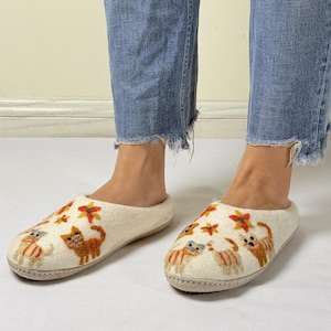 Op de afbeelding: Witte wollen pantoffels met geborduurde katten en bloemen. De pantoffels hebben een afgeronde neus en een platte zool.