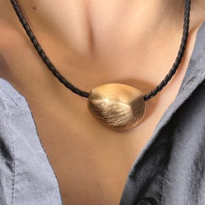 Könnte beinhalten: Eine goldfarbene Anhängerkette mit einem strukturierten, organisch geformten Stein. Der Anhänger hängt an einem schwarzen, geflochtenen Lederband. Die Kette wird über einem grauen Hemd getragen.