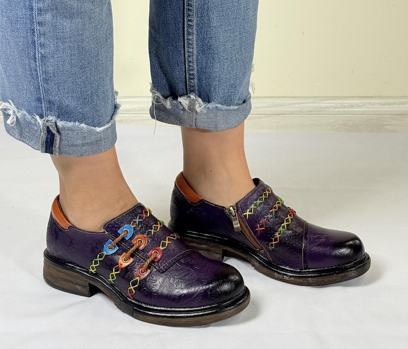 May include: Purple leather shoes with colorful stitching and a zipper detail. The shoes have a chunky heel and a rounded toe.