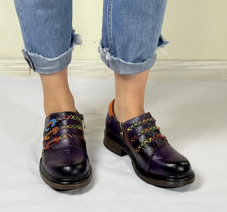 May include: Purple leather shoes with colorful stitching and a zipper detail. The shoes have a chunky heel and a rounded toe.