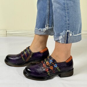 May include: Purple leather shoes with colorful stitching and buckles. The shoes have a chunky heel and a rounded toe.