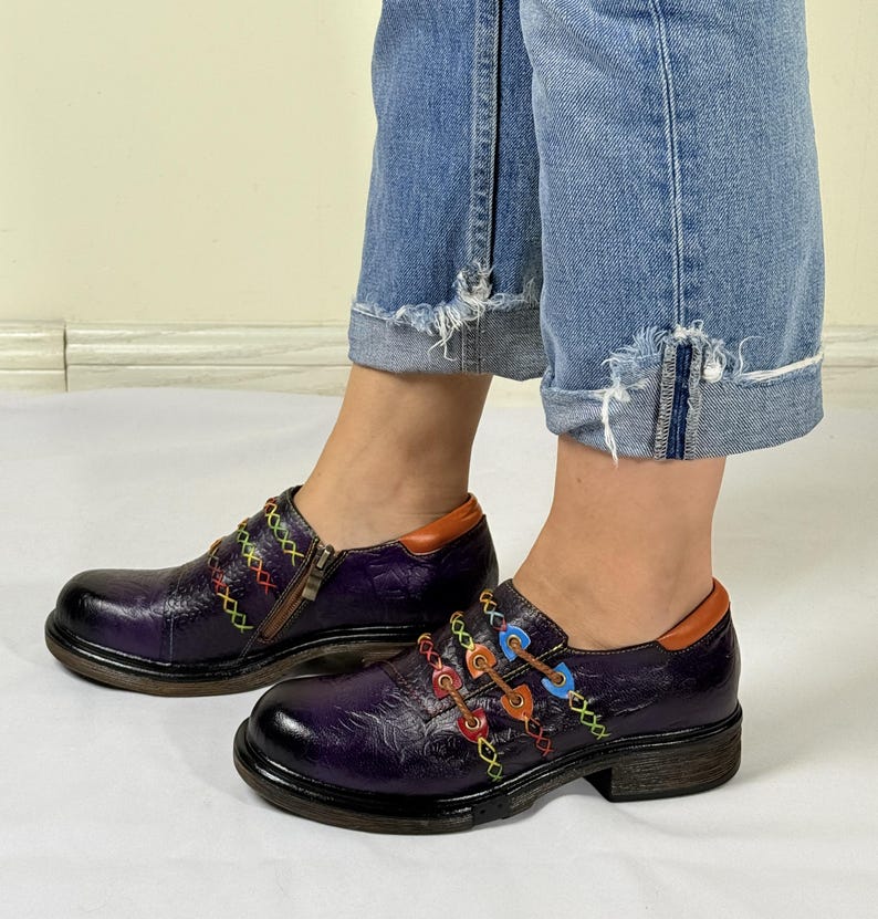 May include: Purple leather shoes with colorful stitching and buckles. The shoes have a chunky heel and a rounded toe.
