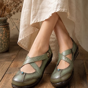 May include: Sage green leather shoes featuring a cross-strap design with embossed floral patterns, a small turquoise-colored stone accent on the toe, and a metallic button detail on the side, set on a dark brown sole with a low heel.
