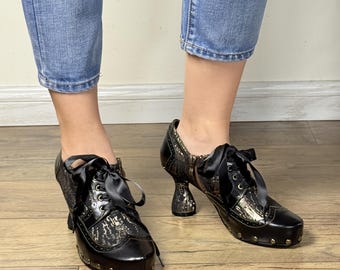 May include: Black and gold platform shoes with a unique heel design. The shoes feature a lace-up closure with black satin ribbons and decorative gold studs along the sole. The upper has a patterned design.
