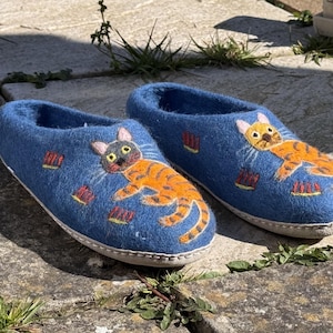 May include: A pair of blue felt slippers with embroidered orange and black cats. The cats are depicted in a playful pose, with one cat on each slipper.