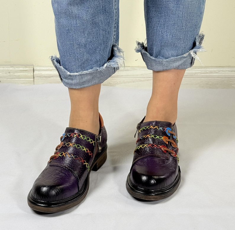 May include: Purple leather shoes with colorful stitching and a zipper detail. The shoes have a chunky heel and a rounded toe.