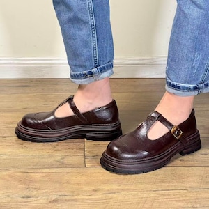 May include: A pair of dark burgundy T-strap shoes with a chunky sole. The shoes have a rounded toe and a buckle closure. The soles are approximately 1.5 inches thick. The shoes are worn with blue jeans.