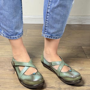 May include: A pair of sage green leather shoes featuring a cross-strap design with embossed floral patterns, a small round turquoise-colored stone accent on the toe, and a metallic button detail on the side strap, worn with blue denim jeans.