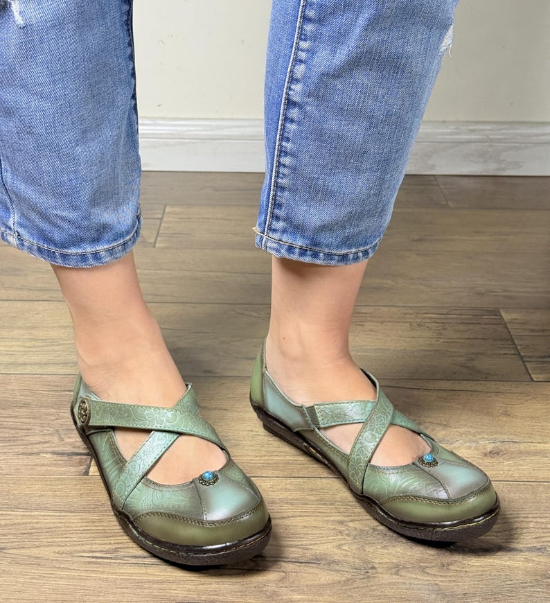 May include: A pair of sage green leather shoes featuring a cross-strap design with embossed floral patterns, a small round turquoise-colored stone accent on the toe, and a metallic button detail on the side strap, worn with blue denim jeans.