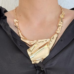 May include: A gold-toned statement necklace with an abstract, geometric design. The necklace features a series of interconnected, folded metal shapes that create a bold and modern look. The necklace is worn against a black shirt.