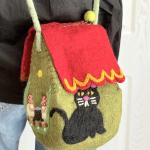 May include: A handmade felt crossbody bag shaped like a house. It has a red roof, a black cat design, and a green body with mushroom and flower details. The strap is light green. Measurements are in centimetres.
